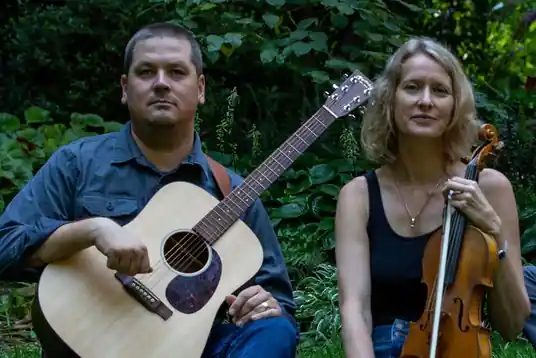 Alex Caton with a fiddle and Jordan Perry with a guitar in a green orchard